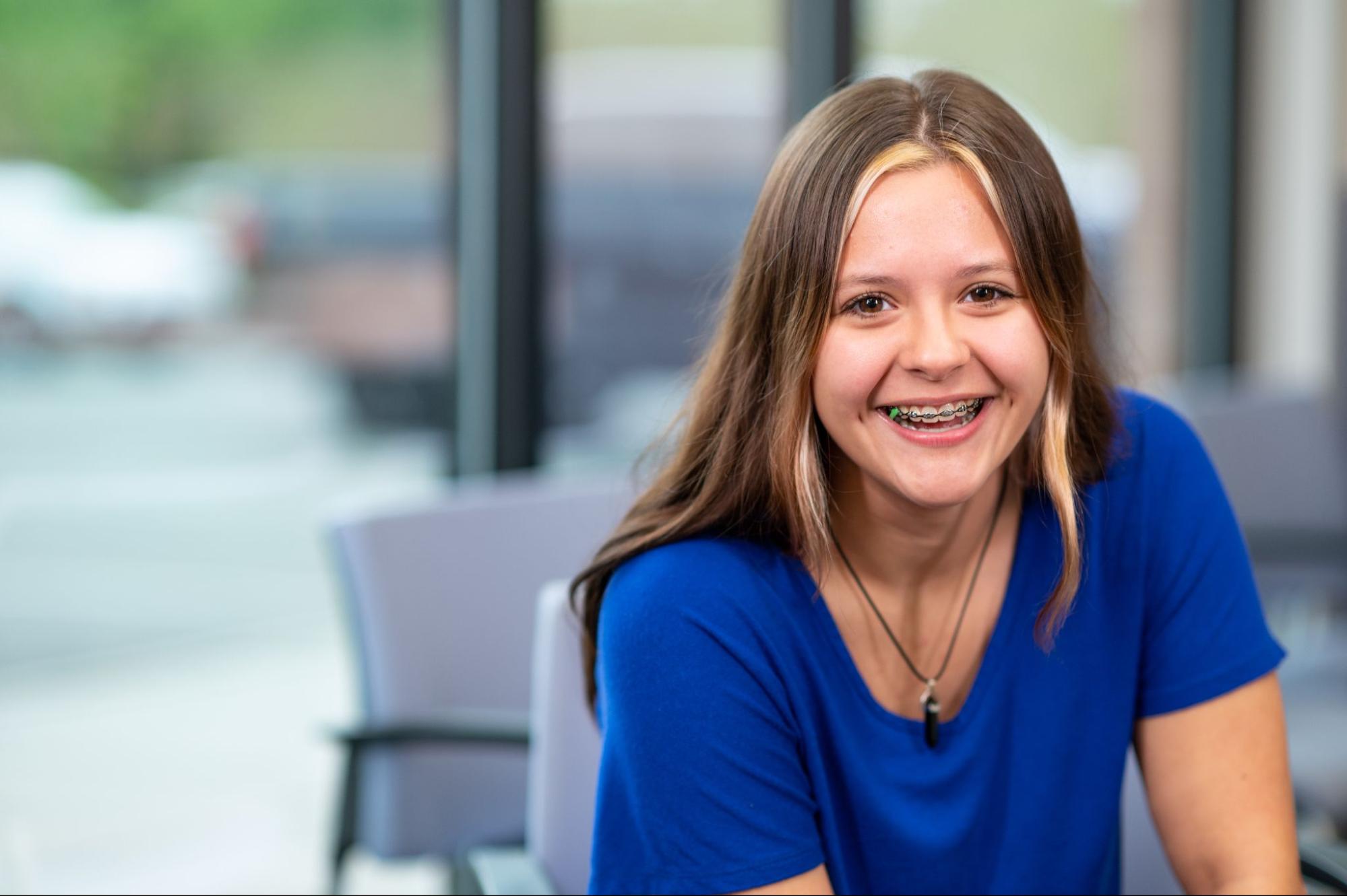 Smiling young girl with braces in blue shirt, sitting in a welcoming orthodontic office environment, reflecting a stress-free orthodontic experience.