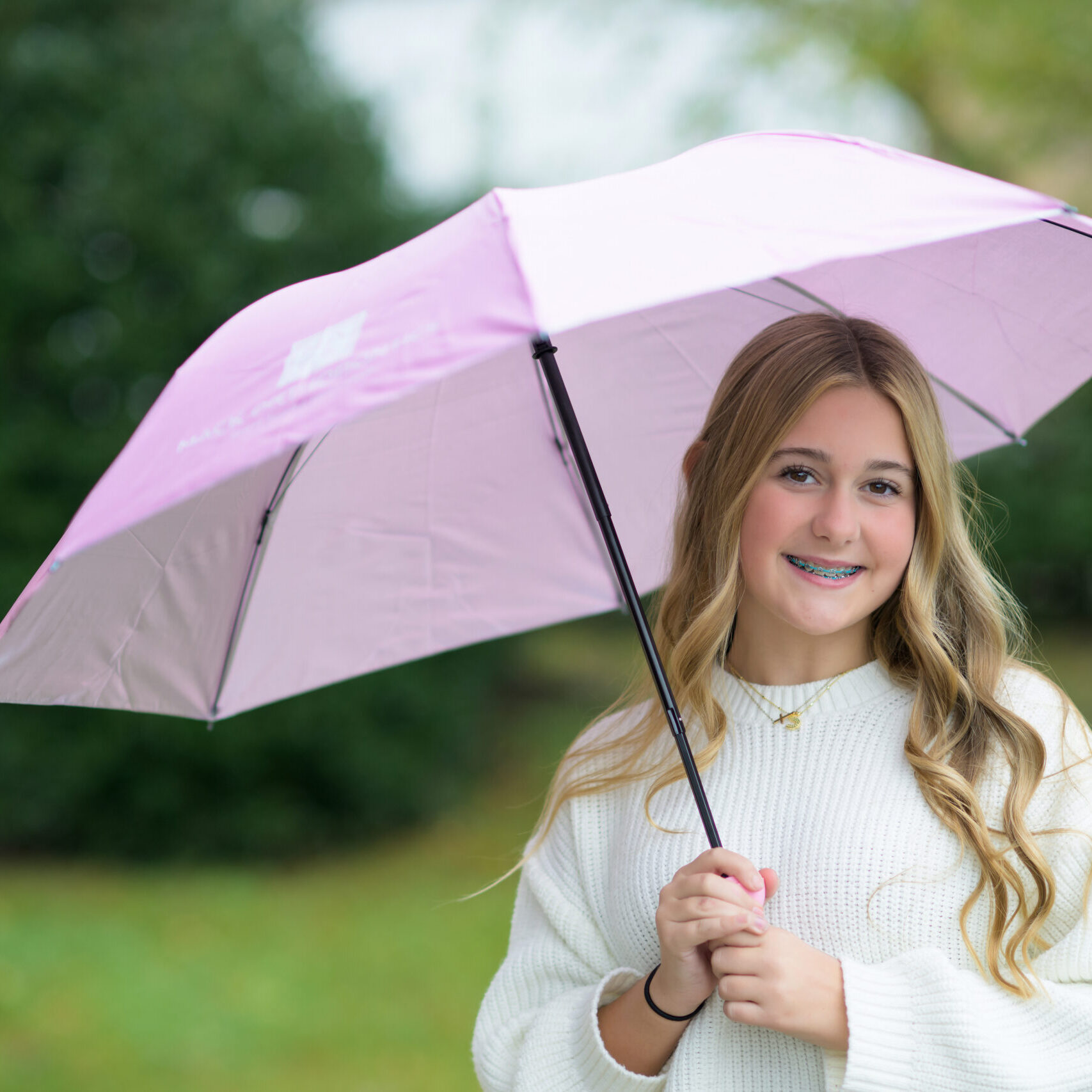 Girl with braces holding a pink umbrella, smiling, in a natural outdoor setting, promoting Mack Orthodontics and inviting inquiries for orthodontic services in Burlington and Hillsborough, NC.